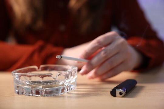 Woman flicking ash from cigarette into ashtray at wooden table indoors, closeup