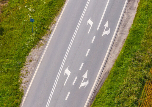 Aerial view of an asphalt road with white directional arrows and lane markings flanked by green grassy verges in Paslek, Warmian-Masurian Voivodeship, Poland.