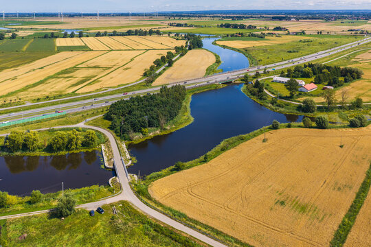 Aerial view of Droga Ekspresowa S7 crossing the landscape with a winding river, golden wheat fields, and wind turbines in the distance in Nowy Dwor Gdanski, Pomeranian Voivodeship, Poland.