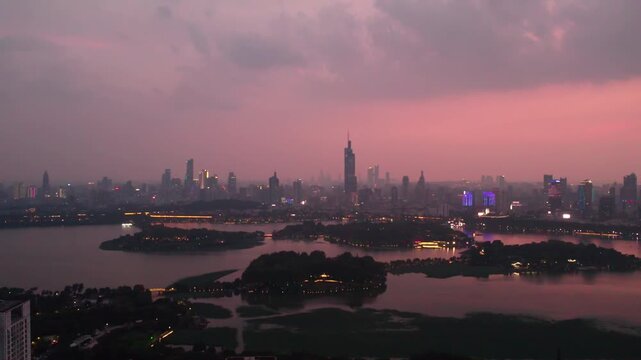 A panoramic dusk view of a city skyline across a calm lake, with warm pink sky glow and lit urban structures.