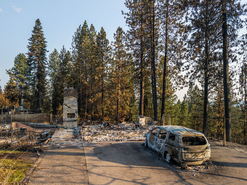Aerial view of wildfire damage showing a destroyed home with a standing stone chimney, a burnt-out vehicle, and scorched pine trees in a forest setting in Spokane, Washington, United States.