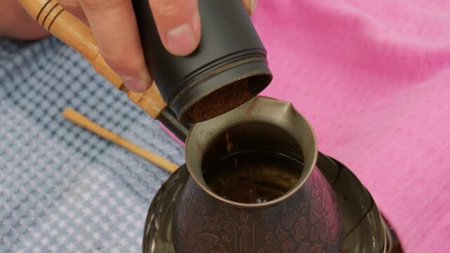 Hand pours ground coffee powder from a container into a traditional metal cezve pot with water. Concept of artisanal coffee brewing, morning ritual, and authentic drink preparation.