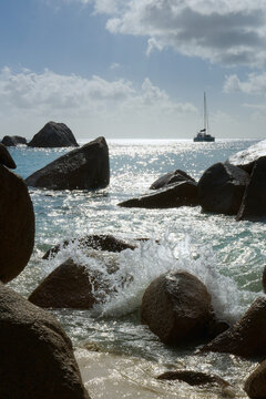 Waves splash against massive granite rocks on a tropical Seychelles coast, with a sailboat on the sparkling turquoise sea under a cloudy sky.
