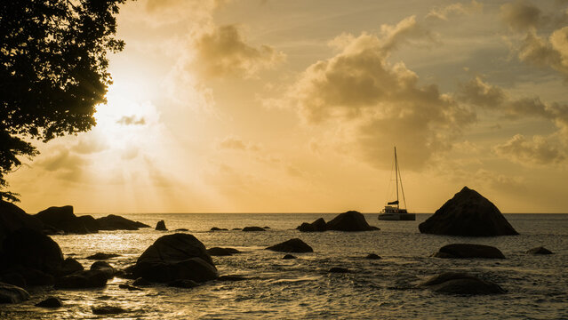 Golden sunset over a tropical Seychelles coastline with granite rocks, calm ocean water and a sailboat silhouetted against the glowing sky.