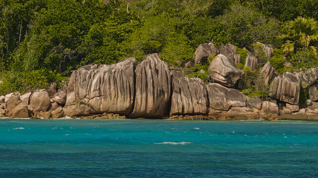 Tropical coastline with massive granite rocks, lush green vegetation and clear turquoise ocean water in the Seychelles.