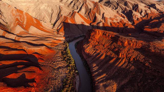 Aerial view of a winding river flowing through deep red rock canyons with dramatic shadows and desert vegetation in Moab, Utah, United States.