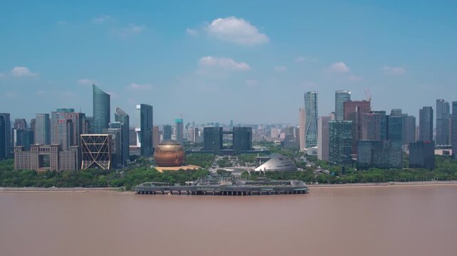 A wide view of a modern city skyline with various skyscrapers and buildings, set along a muddy river under a clear blue sky with scattered clouds.