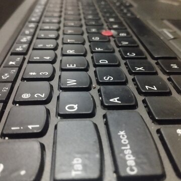 A close-up, low-angle perspective shot of a black laptop keyboard, highlighting the textured keys, white lettering (QWERTY layout), and the iconic red trackpoint button located in the center.