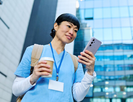 Nurse in scrubs enjoying coffee and using a mobile phone outside a modern medical facility during a sunny day