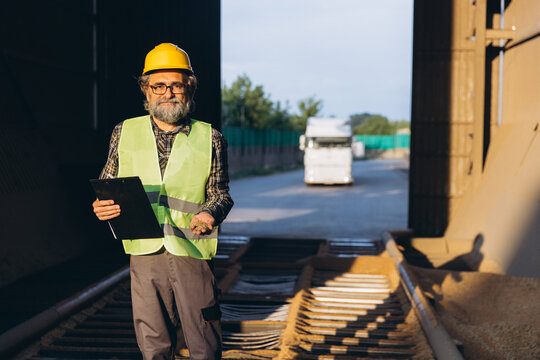 Man in safety vest and hard hat holding a clipboard and handful of grain, supervising a granary elevator with truck