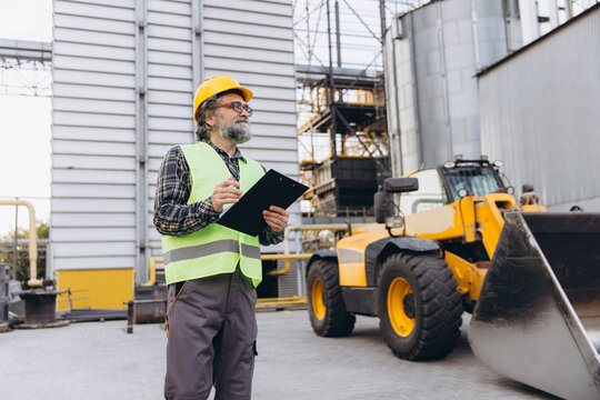 Experienced agricultural engineer inspecting grain elevator facility, writing notes on clipboard with tractor loader parked nearby