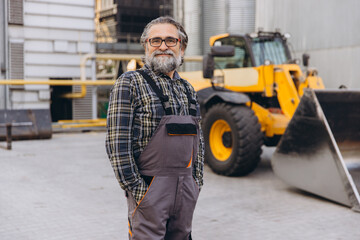 Fototapeta premium Experienced man smiling while standing in front of heavy agricultural machinery at a grain elevator or industrial facility