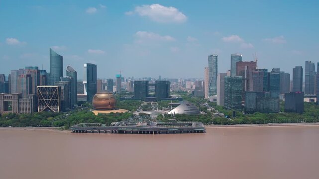A panoramic view of a modern urban skyline along a brown river, with green spaces and distinctive buildings under a clear blue sky.