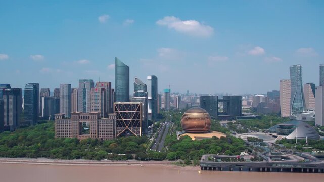 A modern urban skyline with diverse buildings lines a wide brown river under a clear blue sky with scattered clouds.