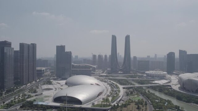A hazy daytime cityscape featuring modern architectural complexes, twin skyscrapers, domed venues, and a winding river in an urban district.