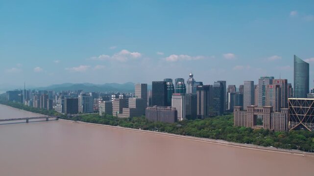 A wide brown river flows past a modern city skyline with dense high-rise buildings, green riverbank trees, distant mountains under a clear blue sky.