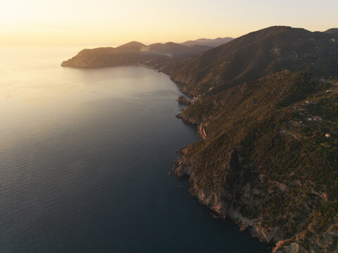 Aerial view of the rugged coastline of Parco Nazionale delle Cinque Terre with steep cliffs, lush vegetation, and calm sea illuminated by golden hour light in Liguria, Italy.