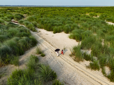 Aerial view of two people walking along a sandy path through tall green grass in a rural landscape in Rajbari, Dhaka Division, Bangladesh.