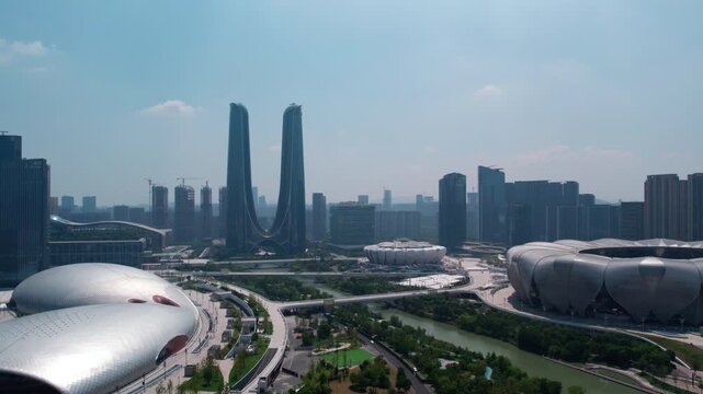 A modern urban panorama featuring twin skyscrapers, large stadiums, a river, and green spaces under a clear sky.