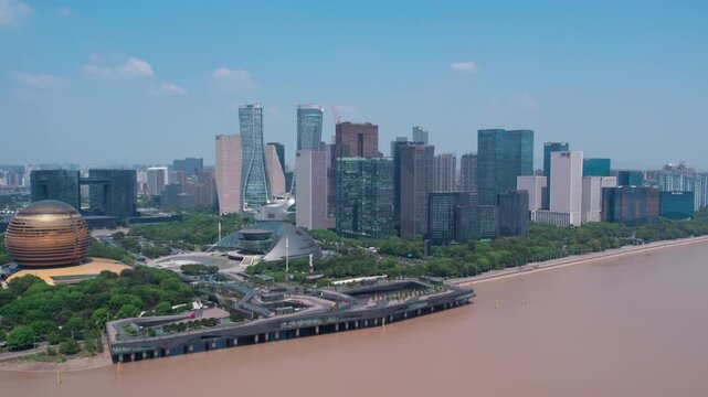 Aerial view of modern urban skyscrapers lining a muddy riverbank, with green spaces under a clear blue sky.