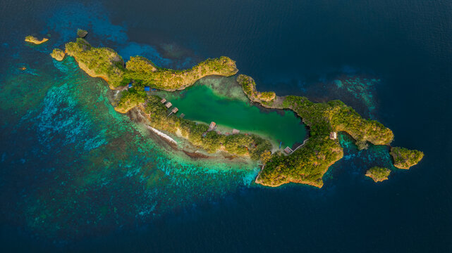 Aerial view of Raja Ampat Regency, a lush tropical island with a turquoise lagoon, dense vegetation, and small wooden huts surrounded by deep blue ocean waters in Southwest Papua, Indonesia.