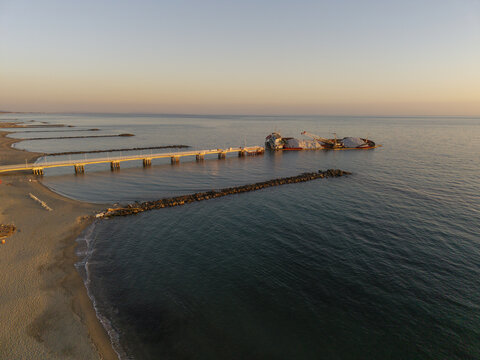 Aerial view of a pier extending into the calm sea at sunset with a cargo ship anchored nearby, sandy beach and breakwaters in Marina di Massa, Tuscany, Italy.