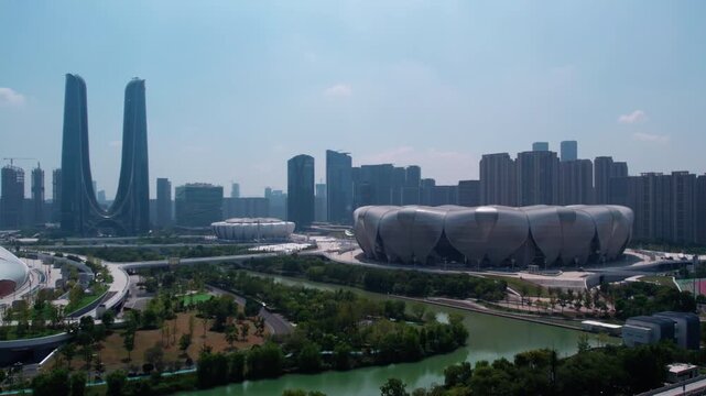 A daytime city view featuring a large modern stadium, twin skyscrapers, urban buildings, a river and green spaces.