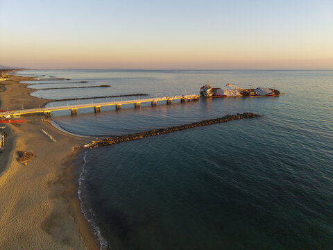 Aerial view of the pier at Marina di Massa with a cargo ship anchored near the breakwaters along the sandy coastline during golden hour in Marina di Massa, Tuscany, Italy.