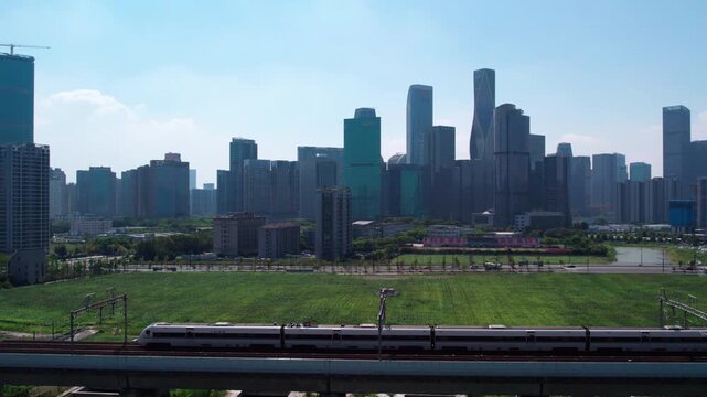 A high-speed train travels on an elevated track, with a modern city skyline and green open land in the background on a clear sunny day.