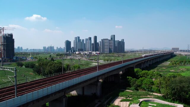 A high-speed train runs on an elevated railway viaduct, with modern urban buildings and green landscapes under a clear sky.