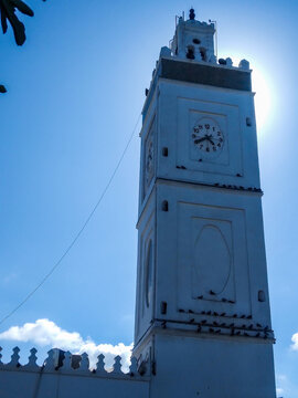 Majestic white minaret clock tower featuring intricate architectural details and dark birds perched against a brilliant bright blue sky captured during a sunny afternoon outdoor session.