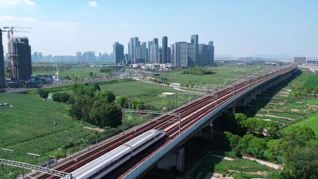 An elevated railway runs through green open land, with modern urban high-rise buildings in the distance under clear sky.