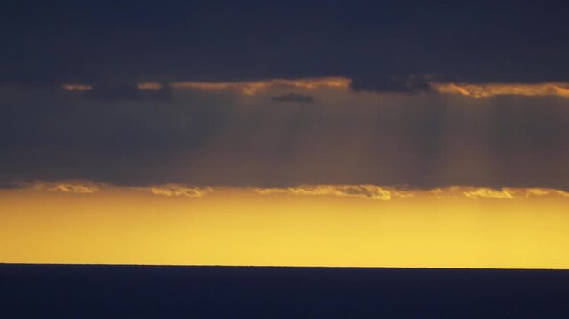 Golden sunset sky illuminates deep shadows over a calm dark sea horizon during evening hours. Static long shot.