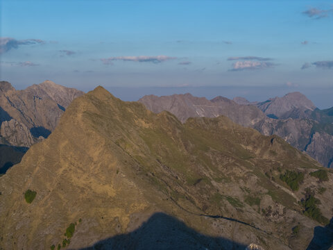 Aerial view of the Apuan Alps mountain range with rugged rocky peaks, golden hour sunlight, and deep shadows under a clear blue sky in Tuscany, Italy.