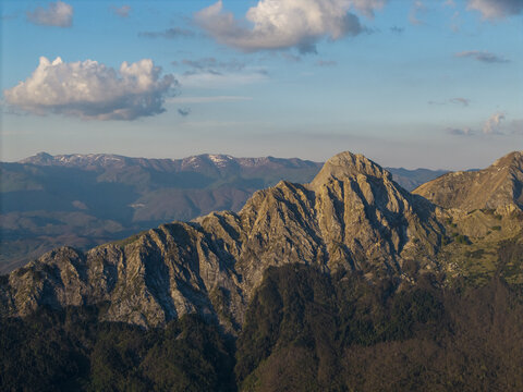 Aerial view of the rugged peaks and rocky slopes of the Apuan Alps under a clear blue sky with scattered clouds in Tuscany, Italy.