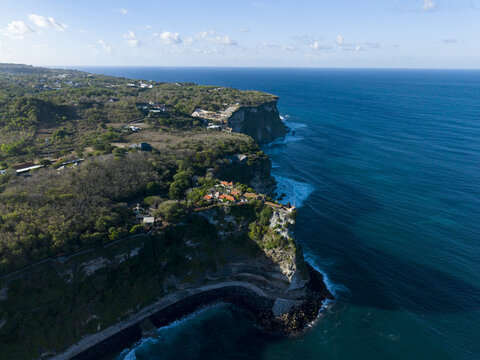 Aerial view of Uluwatu Temple perched on a high cliff overlooking the Indian Ocean, with lush greenery, rocky coastline, and clear blue water in South Kuta, Bali, Indonesia.