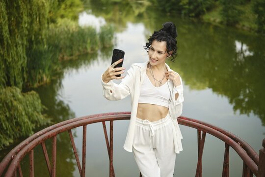 Woman in loose white clothes stands on red bridge over calm water in park and making video call