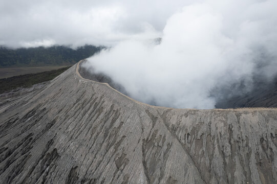 Aerial view of the volcanic crater of Mount Bromo with thick white smoke rising from the rugged, ash-covered slopes under a cloudy sky in Tosari, East Java, Indonesia.