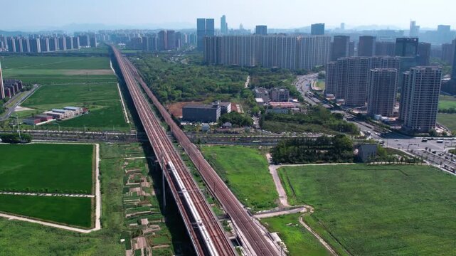 A high-speed train runs on an elevated railway, with urban buildings and green fields in the surrounding landscape.