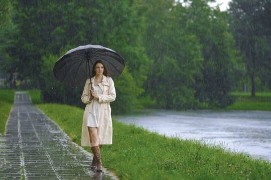 Woman hurries along wet path next to river while holding large umbrella. Rain falls steadily all around as she moves through the scene. Green trees are visible in background