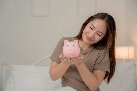 Saving money, financial planning and future goals concept. Happy young Asian woman holds a pink piggy bank at home. Savings, finance, investment, budget, future