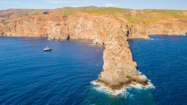Aerial view of Socorro Island featuring rugged cliffs and rocky formations extending into the deep blue ocean with a small boat nearby in Colima, Mexico.