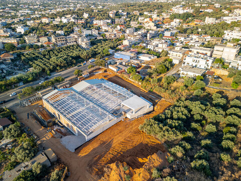 Aerial view of a large construction site featuring a metal frame structure surrounded by olive groves and residential buildings in Chania, Greece.