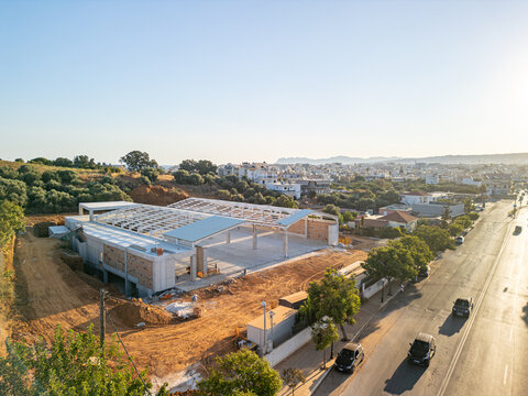 Aerial view of a construction site featuring a steel frame structure on a dirt lot, adjacent to a road with moving cars, with the urban landscape of Chania, Greece.