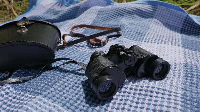 Black vintage binoculars lie on a blue patterned blanket next to their leather carrying case in a sunlit outdoor field. Green grass frames the edge of the picnic setup on a bright day.