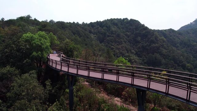 An elevated curved walkway winds through dense green forested mountain slopes in an outdoor natural landscape.