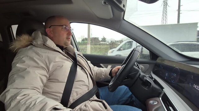 Middle-aged man driving a car on an overcast day while talking. Static interior view focusing on the driver's profile and steering action.