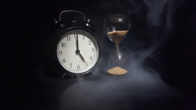 Slow motion vintage alarm showing 5 o'clock and hourglass with falling sand in dramatic smoke on black background. Concept of morning, work time, new day and time management.