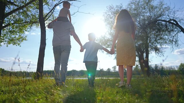 Happy family walks together holding hands near lake. Father carries child on shoulders. Parents and children walk on green grass toward water. Family strolls through park at sunny sunset together.