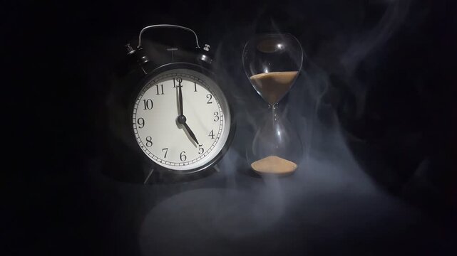 Vintage alarm showing 5 o'clock and hourglass with falling sand in dramatic smoke on black background. Concept of morning, work time, new day and time management.